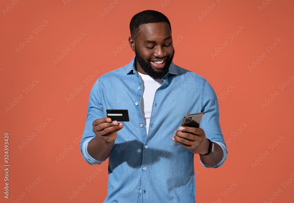 Handsome man makes a payment, using a credit card and smartphone. Photo of african man in casual outfit on coral background. Handsome man makes a payment, using a credit card and smartphone. Photo of african man in casual outfit on coral background.