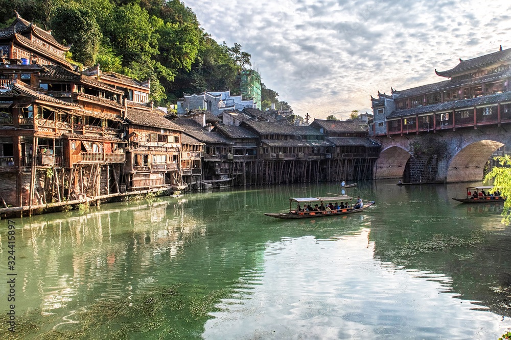 A Small Boat With People Floating Along The River Surrounded By Traditional Chinese Houses (Fenghuang Ancient Town, China)