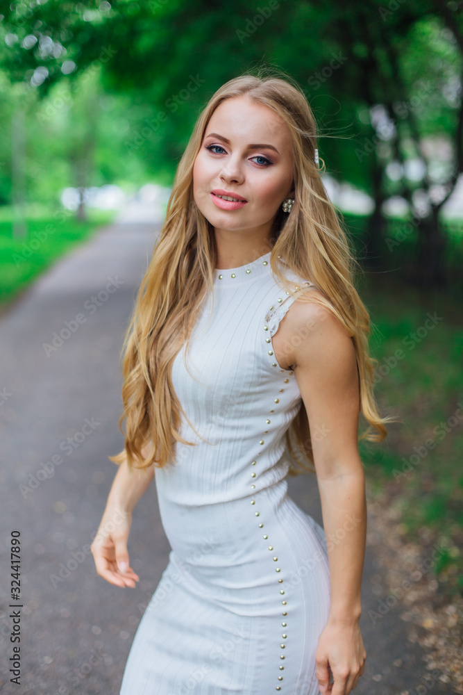 Portrait of a charming blond woman wearing beautiful white dress standing on the road under trees. Portrait of a charming blond woman wearing beautiful white dress standing on the road under trees.