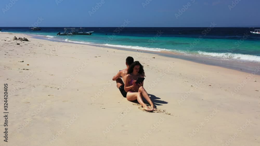Young happy couple sitting back to back on the tropical white sand beach. Woman using tablet and Man reading travel guide. Thailand. Young happy couple sitting back to back on the tropical white sand beach. Woman using tablet and Man reading travel guide. Thailand.