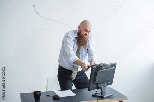 Angry bald man with a red beard in the office in a business suit crashes a computer. Manager with a nervous breakdown breaks the keyboard on the monitor.