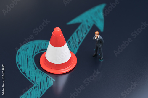 Obstacle, solution idea for business problem or blocker to success concept, miniature people businessman thinking with chalk drawing arrow turn pass the road block traffic pylon on dark blackboard photo