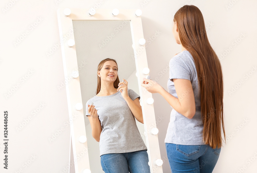 Beautiful young woman applying perfume in front of mirror at home