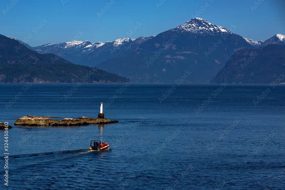 A motor boat carries tourists from Horseshoe Bay to observe whales in the Strait of George,  blue water in a bay against a mountain range and clear blue sky