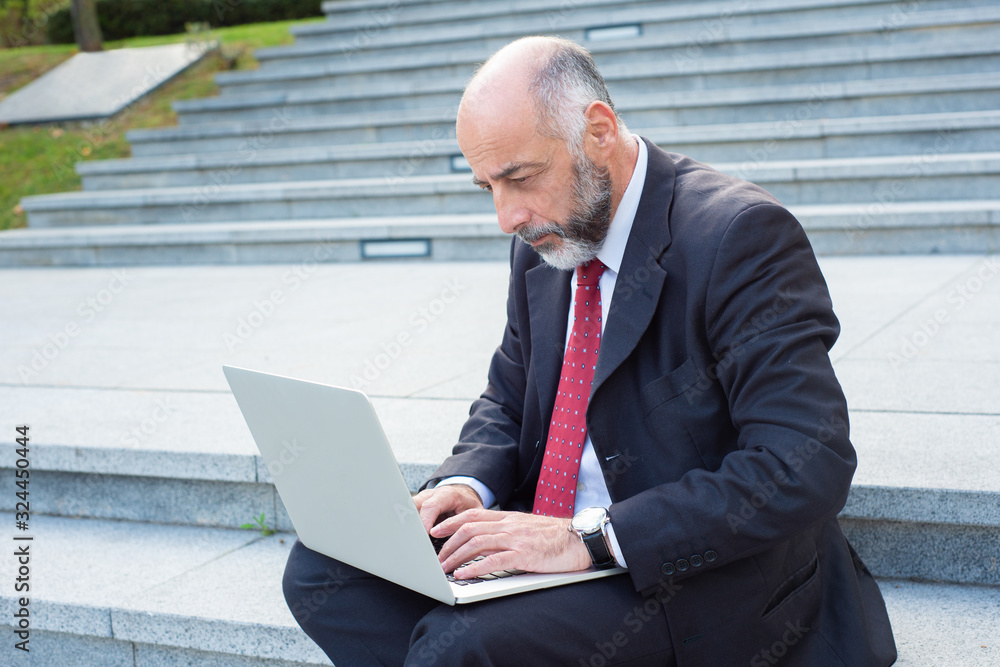 Thoughtful businessman using laptop while sitting on stairs. Focused middle aged attorney typing on laptop outdoors. Technology, business concept Thoughtful businessman using laptop while sitting on stairs. Focused middle aged attorney typing on laptop outdoors. Technology, business concept