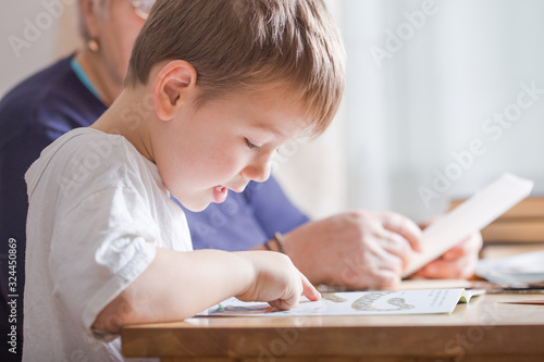 little boy 4 years old reading book. He is sitting on chair in sunny living room watching pictures in story. Kid doing homework for elementary school or kindergarten. Children study.