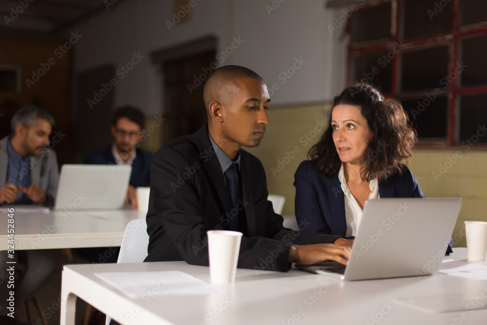 Focused business partners looking at laptop in office. Front view of confident employees talking while looking at laptop. Business, working late concept Focused business partners looking at laptop in office. Front view of confident employees talking while looking at laptop. Business, working late concept