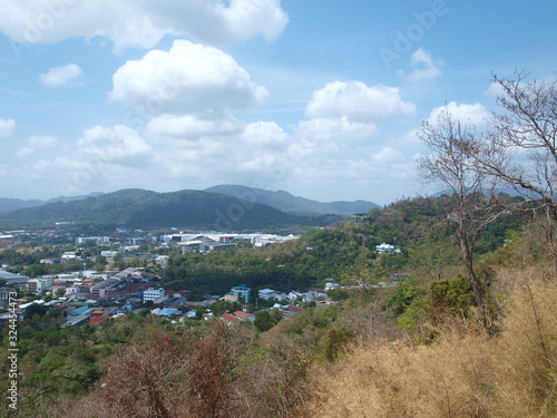 Panoramic view of Phuket Town from a height. Autumn landscape. Yellow  dry and green plants on a foreground  roofs of city buildings in a center  hills and cloudy hills on a background. Cityscape. 