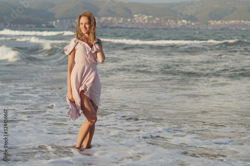 A young girl on the beach in a summer dress stands on the background of the sea, waves and wind, the concept of happiness and relaxation.
