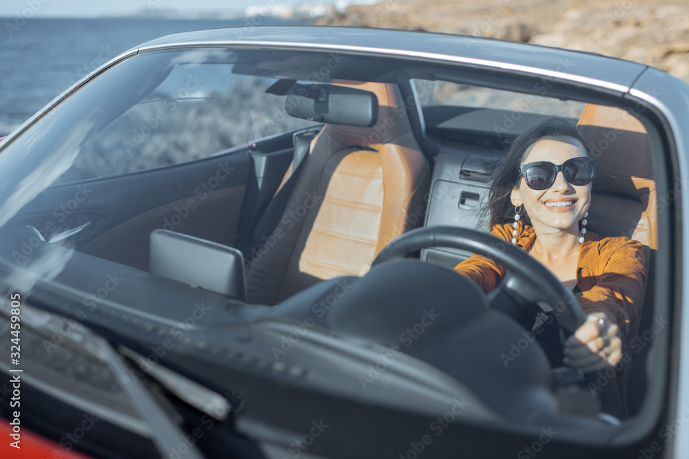 Portrait of a young woman driving a cabriolet near the ocean on a summer time. View through the windshield. Concept of a carefree travel and summer time Portrait of a young woman driving a cabriolet near the ocean on a summer time. View through the windshield. Concept of a carefree travel and summer time