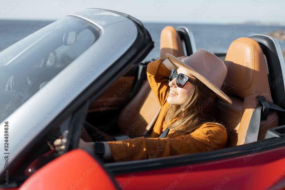Young stylish woman in hat driving convertible car near the ocean, enjoying summer vacations while traveling on the island. Concept of a carefree travel and summer time