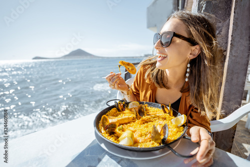 Woman eating paella, traditional spanish dish, while sitting at the restaurant terrace near the ocean. Concept of sea food and good summer vacations