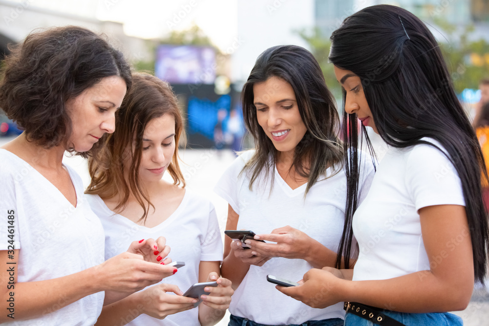 Cheerful women standing in circle and using smartphones. Smiling female friends standing on street with smartphones. Technology concept Cheerful women standing in circle and using smartphones. Smiling female friends standing on street with smartphones. Technology concept