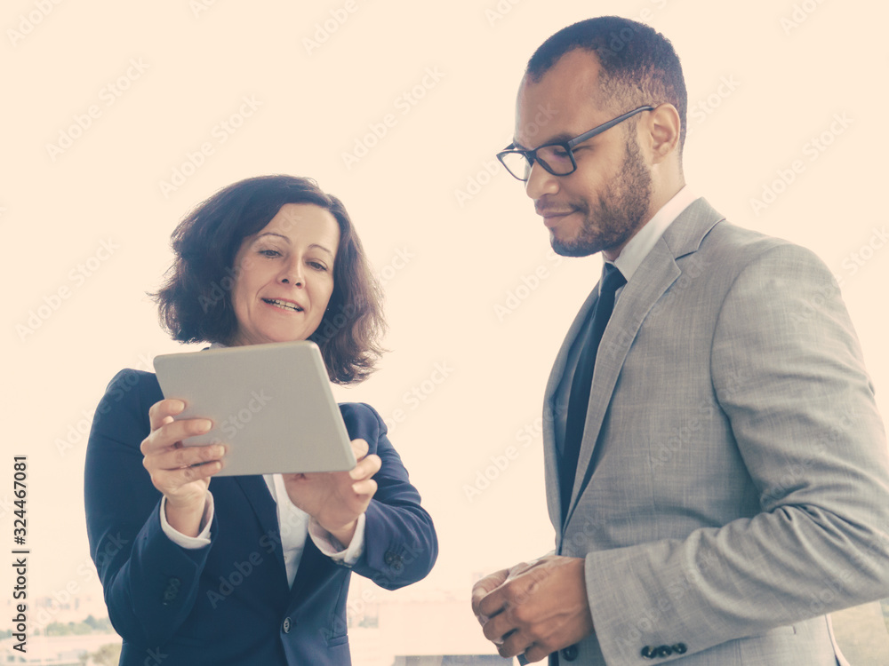 Satisfied business leaders watching project presentation on tablet. Business man and woman standing indoors at office window, woman holding tablet and talking. Business communication concept Satisfied business leaders watching project presentation on tablet. Business man and woman standing indoors at office window, woman holding tablet and talking. Business communication concept