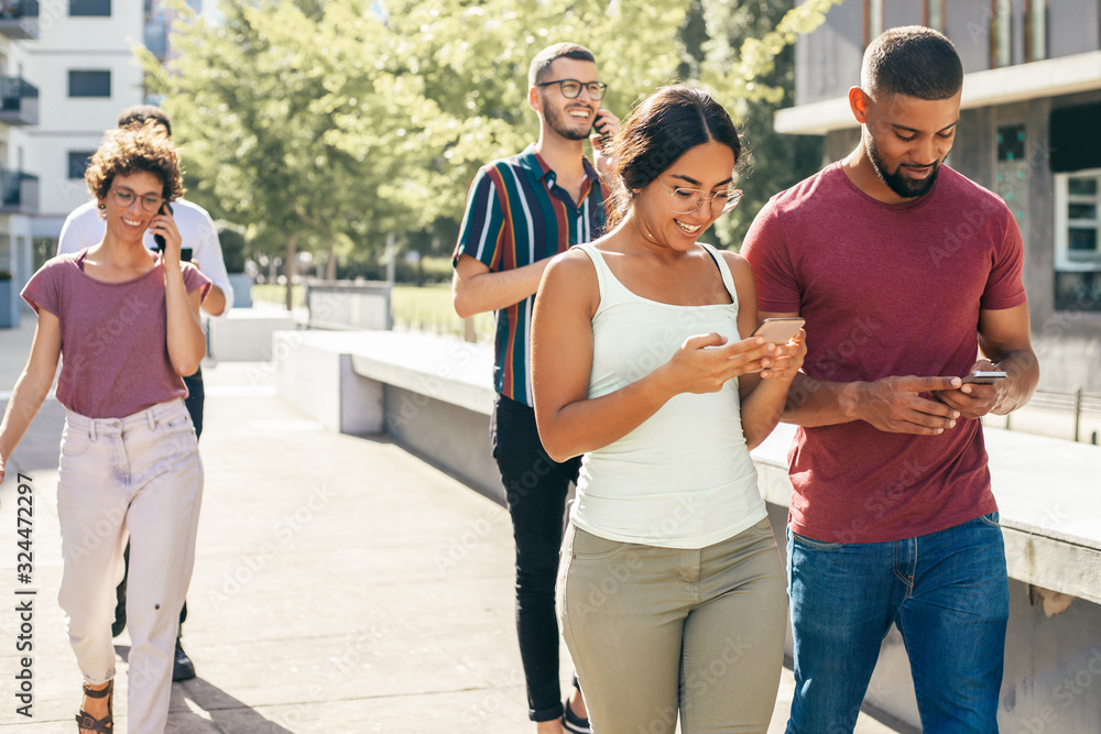 Happy couple using mobile phones outdoors. Young Latin man and woman with smartphones walking outside, people talking on phones behind them. Mobile technology concept
