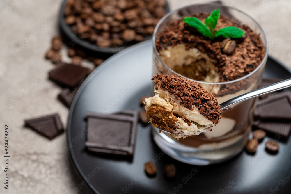 Classic tiramisu dessert in a glass cup, pieces of chocolate on concrete background Classic tiramisu dessert in a glass cup, pieces of chocolate on concrete background