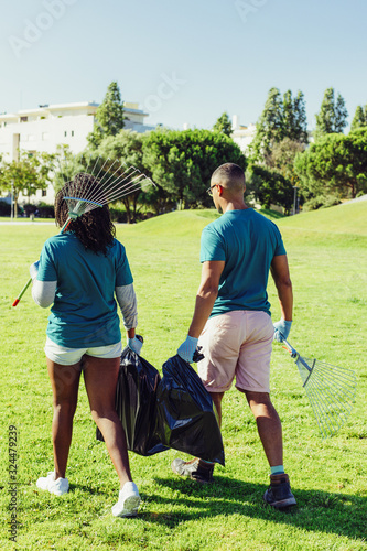 Volunteers cleaning lawn from litter. Rear view of black woman and Latin man walking through grass and carrying rakes and plastic bags. Garbage collection concept