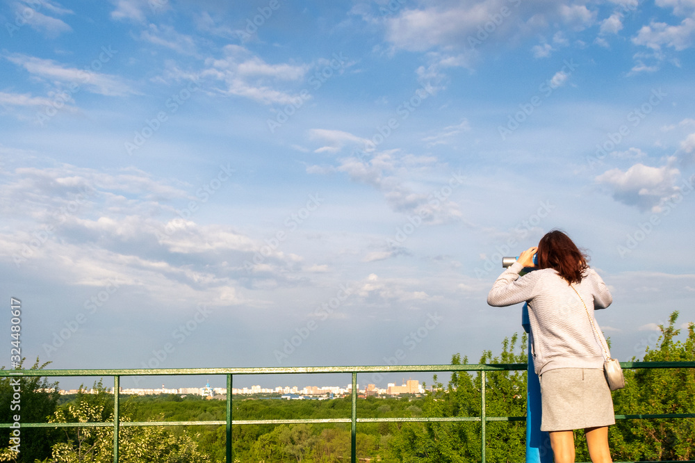 Girl is looking through binoculars at observation deck in public park. Sunny summer day.