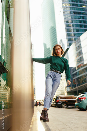 Confident hipster girl with short blond hairstyle, wearing in blue, jeans, boots, posing against rough street wall, minimalist urban clothing style, at sunset background.