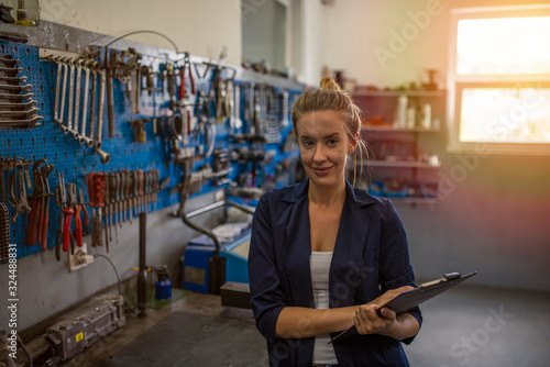 Young woman working in factory. A young female mechanic stands in a garage repair shop. She is wearing blue overalls and smiling to camera. Young beautiful female mechanic posing with clipboard
