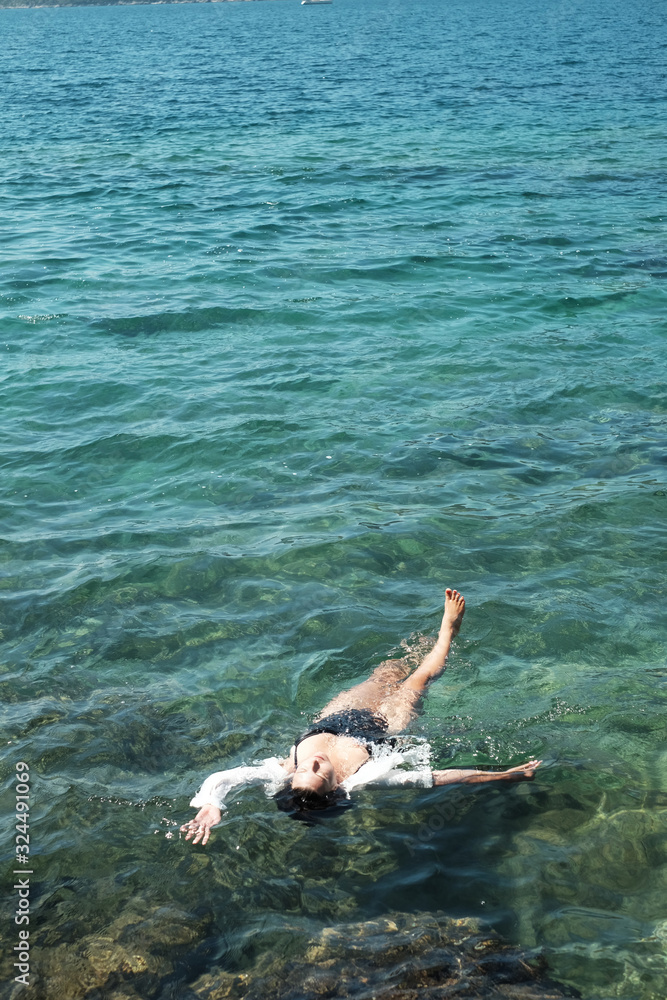 Woman in black swimsuit and white shirt lying on transparent turquoise water surface