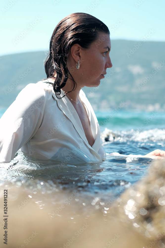 Obraz premium Woman in focus in black bikini and white wet shirt behind unfocused cliff in turquoise water surface. Vertical photo