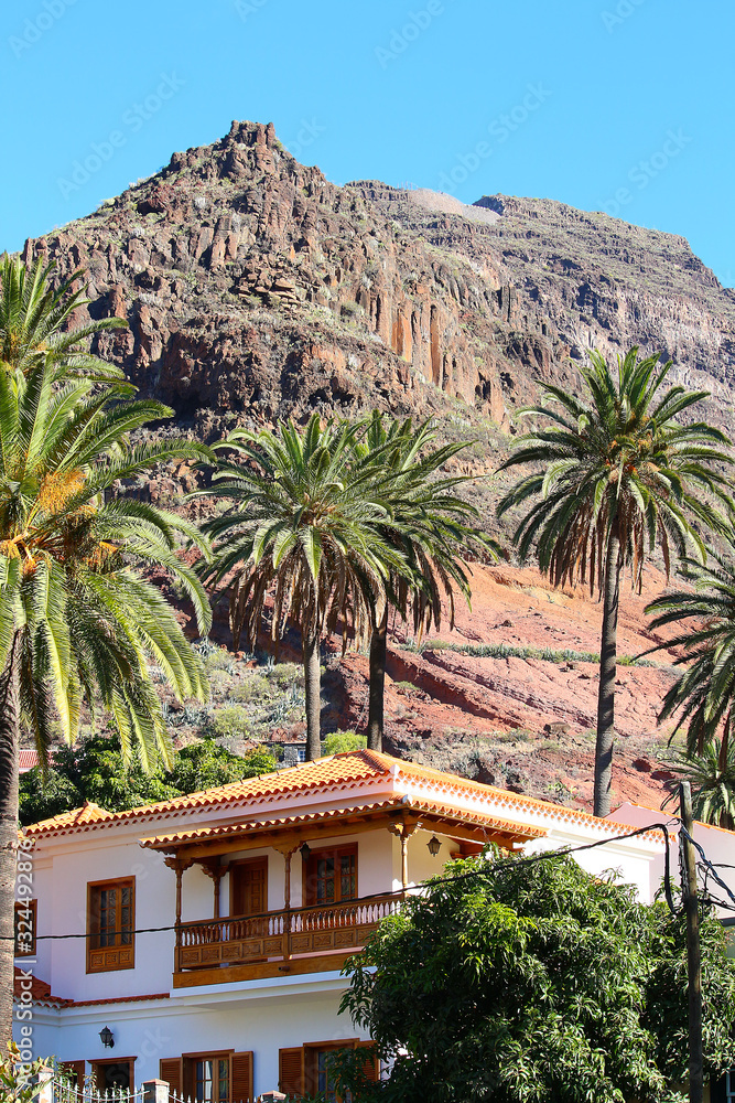 Residential home with woodcrafted balcony balustrade in colonial style in front of mountain and palm trees (La Gomera, Spain)