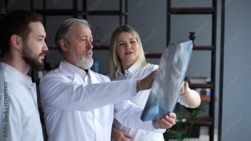 Medical team of three adult and young doctors examining X-ray scan together in the consulting room of the hospital. Concept of team of doctors working together at clinic. Tracking shot in slow motion. Medical team of three adult and young doctors examining X-ray scan together in the consulting room of the hospital. Concept of team of doctors working together at clinic. Tracking shot in slow motion.