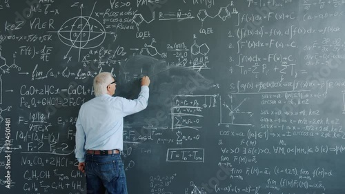 Mature man physicist writing formulas on blackboard with chalk indoors at school busy with solving scientific problem. Science, occupation and education concept.
