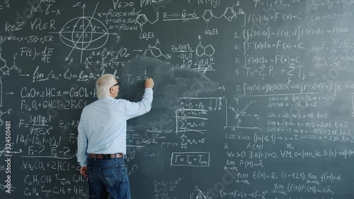 Mature man physicist writing formulas on blackboard with chalk indoors at school busy with solving scientific problem. Science, occupation and education concept. photo