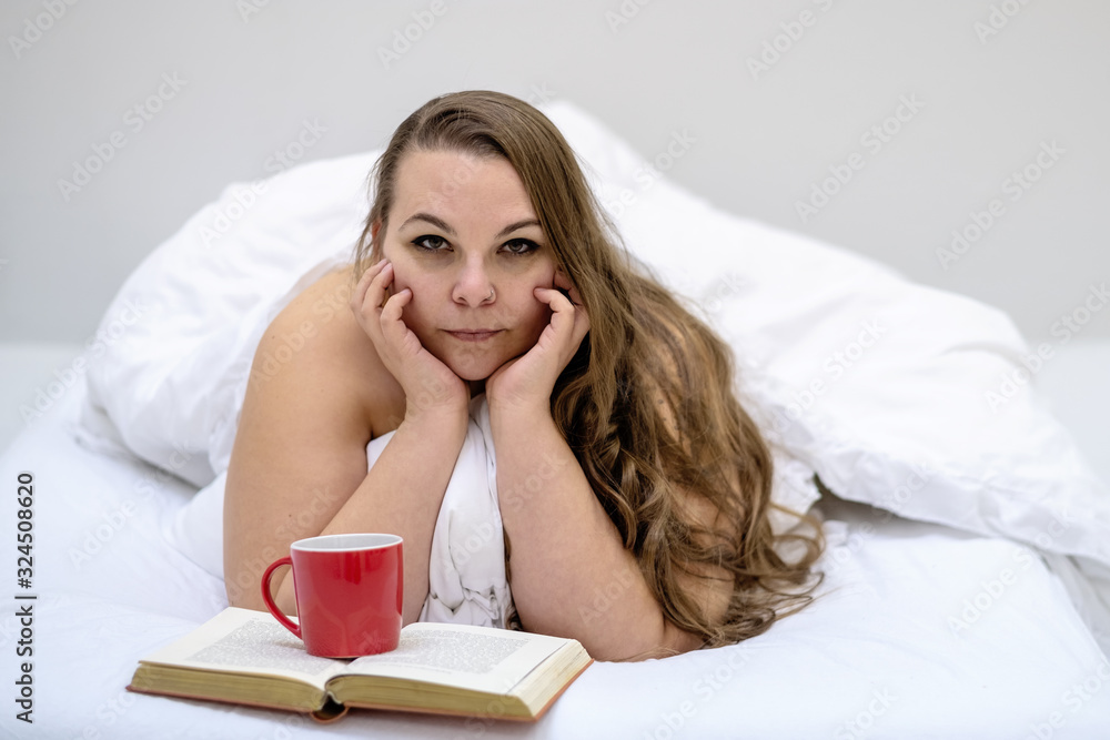 Portrait of an attractive, contented, young, plus size, fat, overweight, chubby, sexy long haired woman lying relaxed in bed enjoys her book and coffee in a red cup in fresh, white bedding. Portrait of an attractive, contented, young, plus size, fat, overweight, chubby, sexy long haired woman lying relaxed in bed enjoys her book and coffee in a red cup in fresh, white bedding.