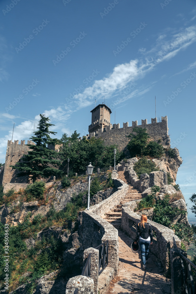 Woman dressed in a motorcycle outfit and sunglasses. body protection turtle and knee pads, helmet in hand. Fortress, stone steps, stairs. Vertical photo. Pass of the witches San Marino