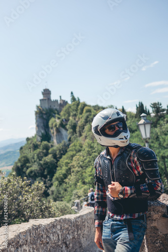 Man dressed in a motorcycle outfit and white helmet and sunglasses. body protection turtle. Fortress on background. Mountains. Stylish. Vertical photo. Pass of the witches San Marino, Italy