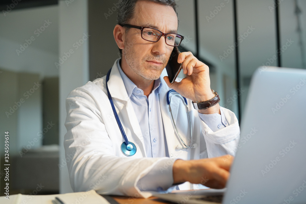 Doctor in office working on laptop talking on phone Doctor in office working on laptop talking on phone