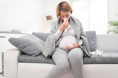 Pregnant woman catching cold, flu, virus, sitting at home on a couch, blowing nose and being sick.