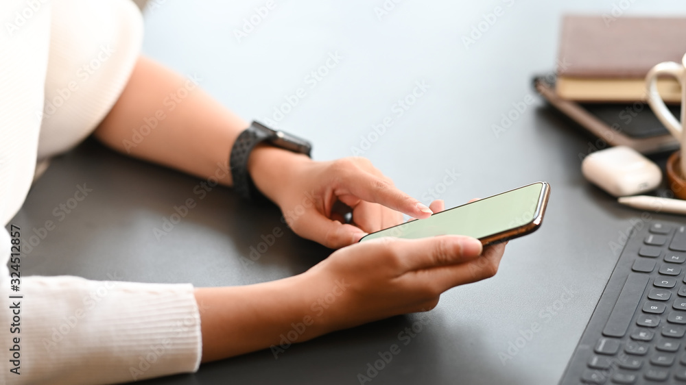 Fototapeta premium Cropped image of creative woman working as graphic designer holding on smartphone while sitting at the modern working table with comfortable living room as background.