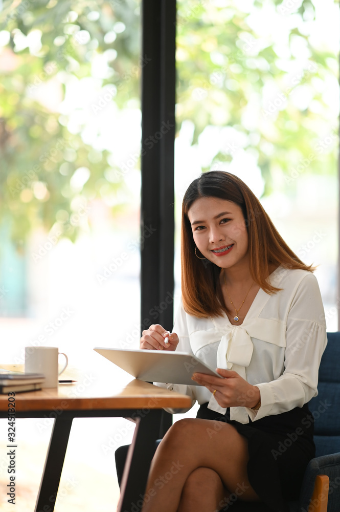 Portrait of young beautiful woman working as secretary using a computer tablet with stylus pen while sitting at the wooden working table with orderly office wall glass as background. Portrait of young beautiful woman working as secretary using a computer tablet with stylus pen while sitting at the wooden working table with orderly office wall glass as background.