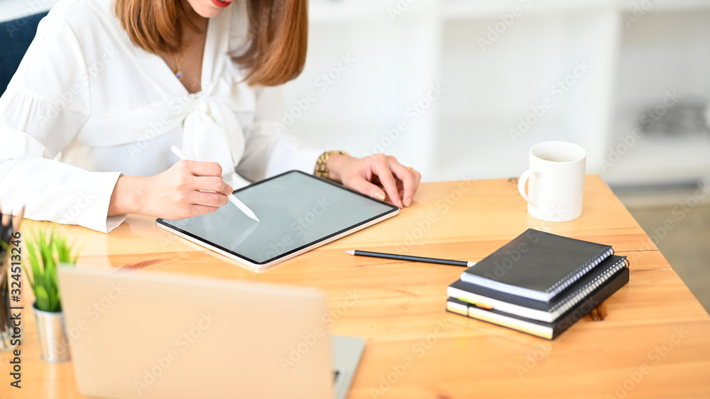 Cropped image waist up of beautiful woman working as secretary drawing a business plan on white blank screen laptop while seated at the modern wooden table with orderly office as background.