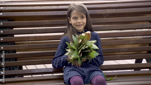 Little beautiful girl in a blue dress with leaves in her hands laughs on a bench in the park