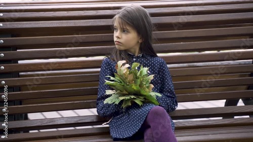 Little beautiful girl in a blue dress with leaves in her hands laughs on a bench in the park