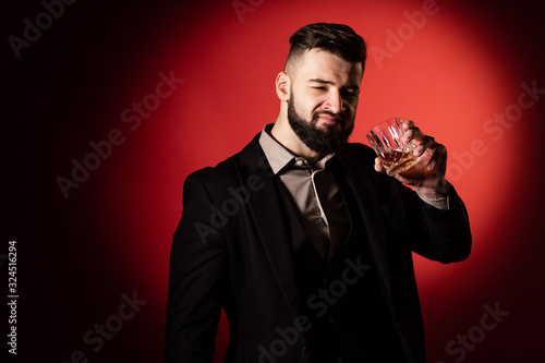 Handsome bearded man in suit with stylish beard holding a glass of whiskey and going to drink it, on red background