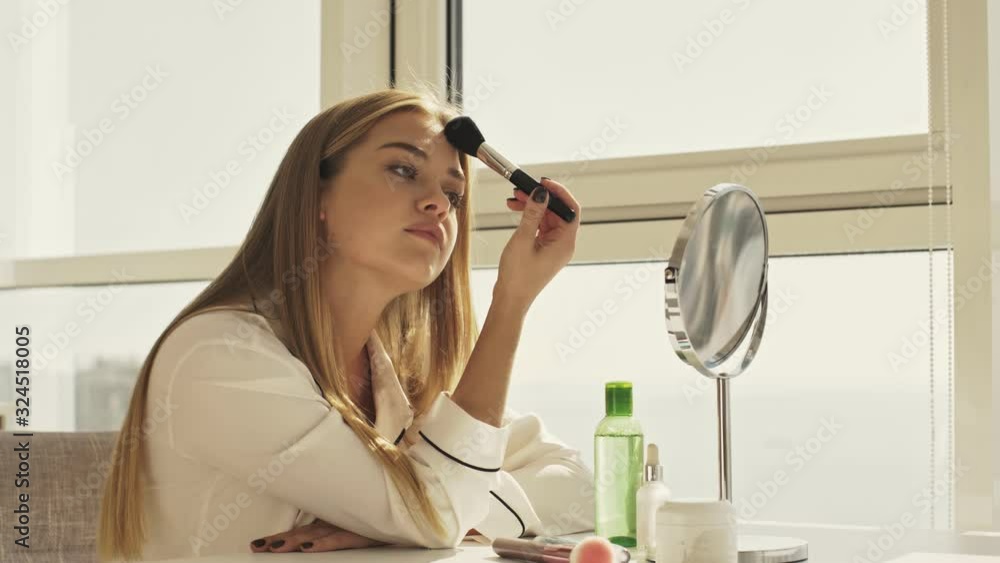 A happy cute woman in pajama is using a powder brush and smiling while sitting at table in bright room
