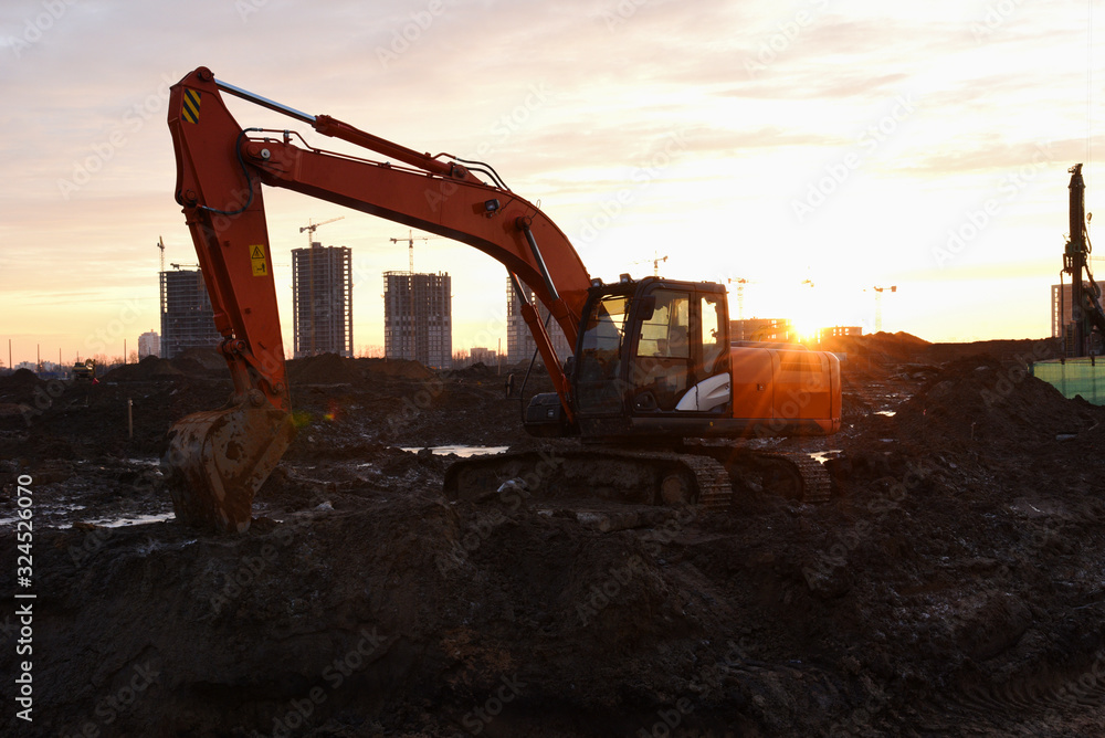 Large tracked excavator at construction site on the background sunset. Backhoe dig the ground for the foundation, laying storm sewer pipes. Installation of water main systems. Road repair Large tracked excavator at construction site on the background sunset. Backhoe dig the ground for the foundation, laying storm sewer pipes. Installation of water main systems. Road repair