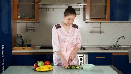 pretty girl in pink dressing-gown makes salad standing at kitchen table and cutting fresh vegetables closeup photo