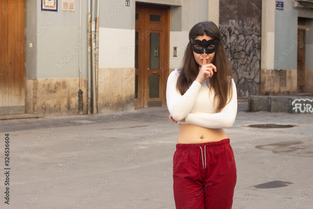 Fototapeta premium Portrait of attractive caucasian young woman with mask holding index finger near his lips, in the city, white top and red pants, dark air. Place for your text in copy space.
