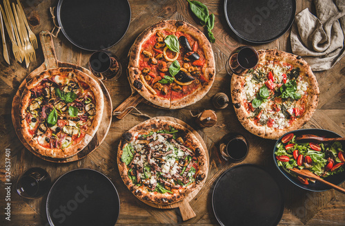 Pizza party dinner. Flat-lay of various kinds of Italian pizza, fresh salad and red wine in glasses over rustic wooden table, top view. Fast food lunch, celebration, gathering concept