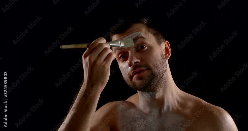 portrait of an unshaven brunette on a black background. he applies white paint to his face with a brush, carefully painting over his eyebrows portrait of an unshaven brunette on a black background. he applies white paint to his face with a brush, carefully painting over his eyebrows