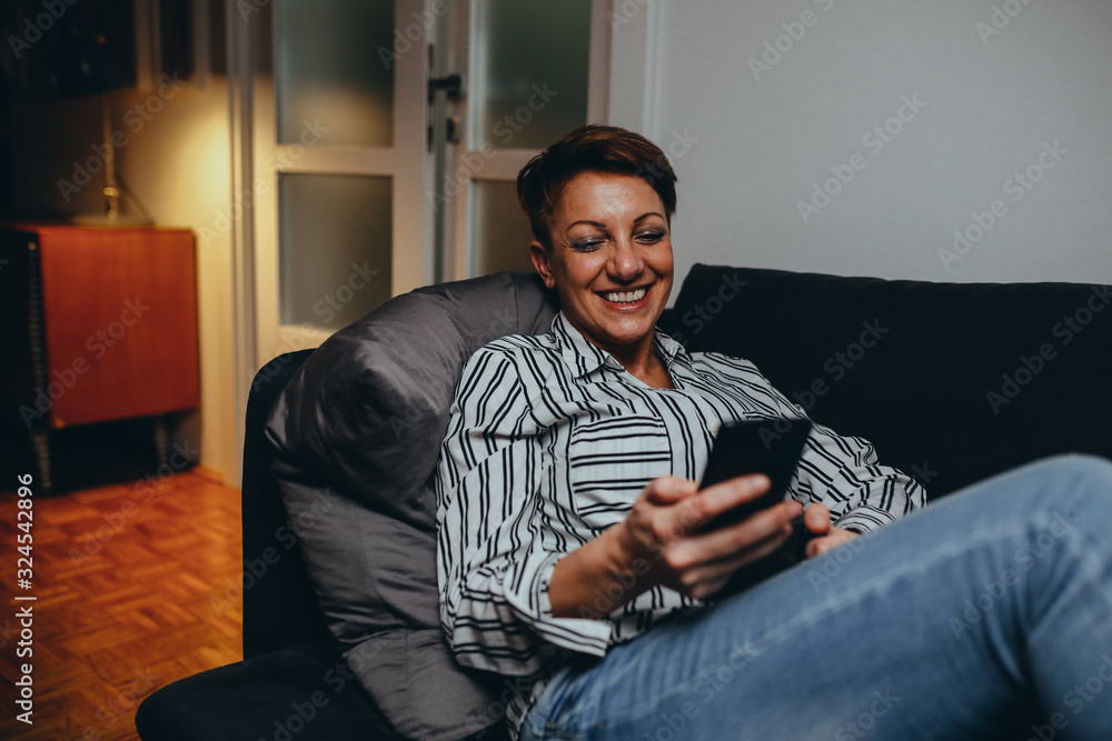 middle aged woman relaxing on sofa , drinking coffee and using mobile phone. moody night scene