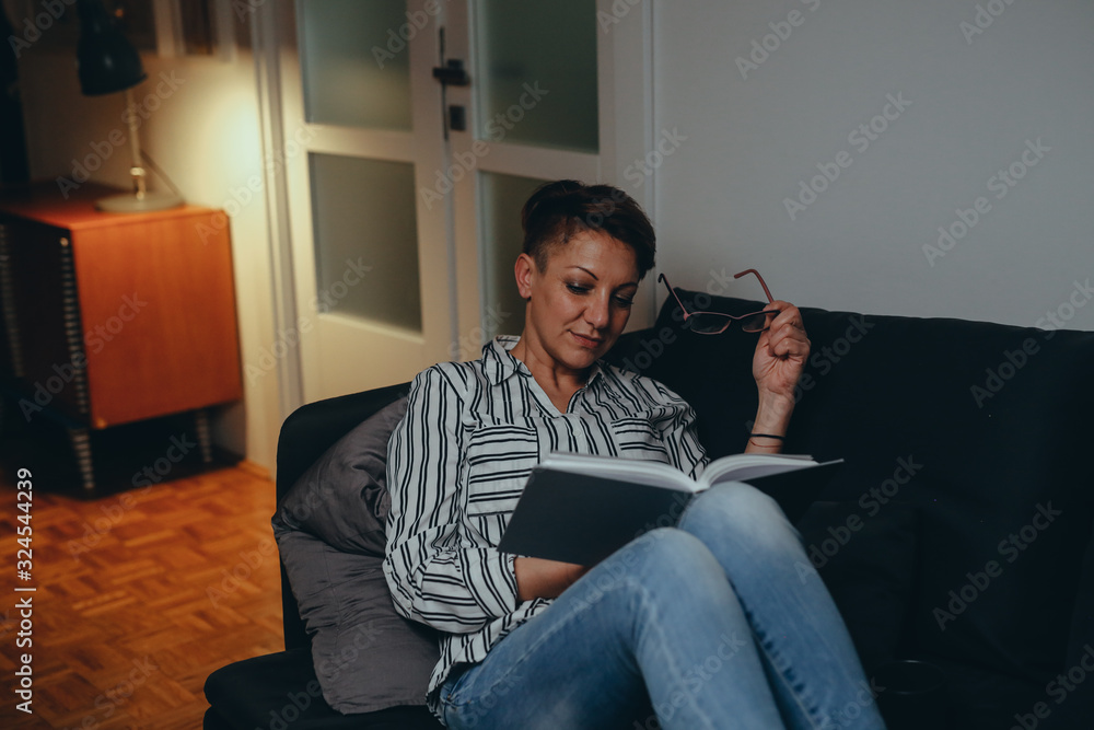 Obraz premium mid aged woman relaxing on sofa and reading book, at her home