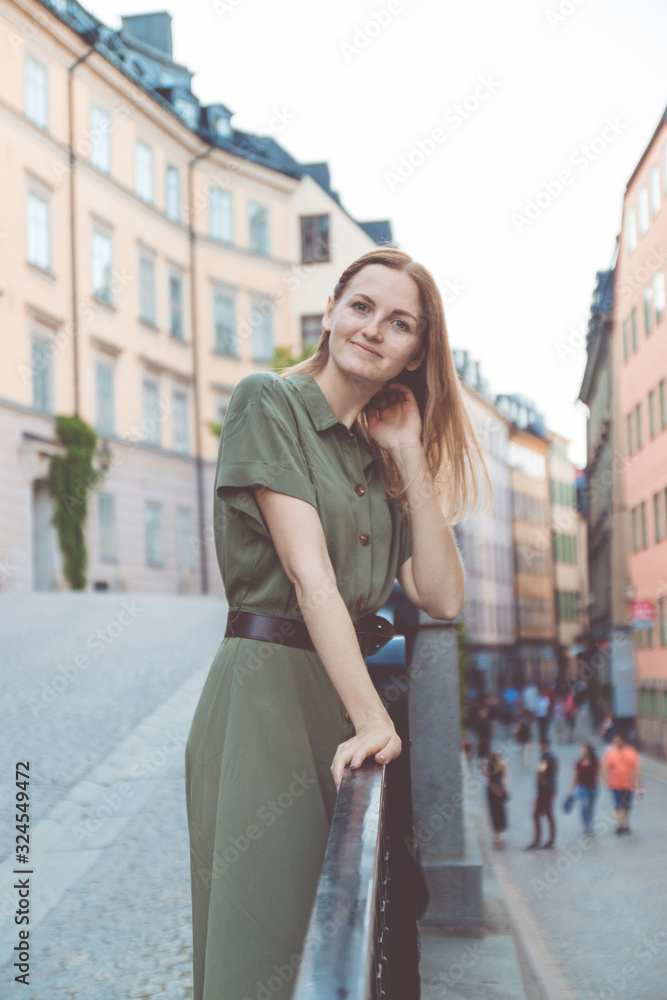 Young blonde in an olive color dress smiles at the camera posing on a city street. Stockholm Old Town, Sweden